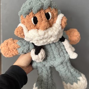 Op de afbeelding: Een gehaakt pluche speelgoed in de vorm van een persoon die een blauwe en witte gebreide muts en een wit gezichtsmasker draagt. Het speelgoed heeft een lichtbruin gezicht met zwarte ogen en een witte baard.