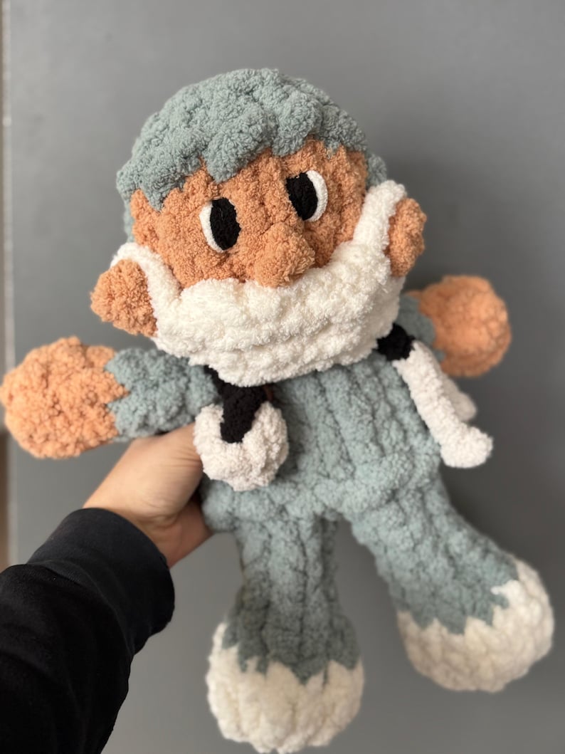 Op de afbeelding: Een gehaakt pluche speelgoed in de vorm van een persoon die een blauwe en witte gebreide muts en een wit gezichtsmasker draagt. Het speelgoed heeft een lichtbruin gezicht met zwarte ogen en een witte baard.