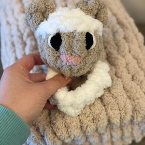 May include: A soft, brown and white knitted baby blanket with a plush, brown and white lamb rattle attached. The blanket has a chunky knit texture.