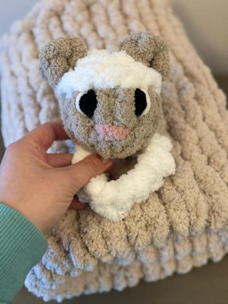May include: A soft, brown and white knitted baby blanket with a plush, brown and white lamb rattle attached. The blanket has a chunky knit texture.