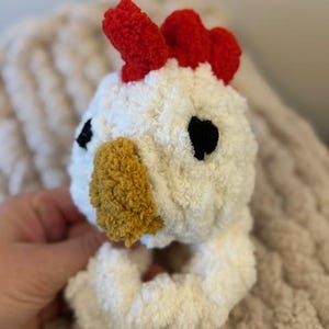 May include: White crocheted chicken toy with a red comb, black eyes, and a yellow beak. The toy is a ring that can be used as a teething toy or a rattle.