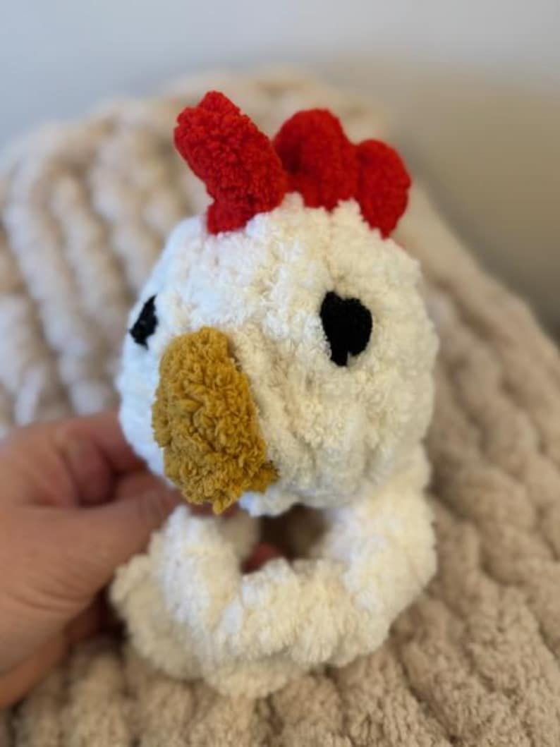 May include: White crocheted chicken toy with a red comb, black eyes, and a yellow beak. The toy is a ring that can be used as a teething toy or a rattle.