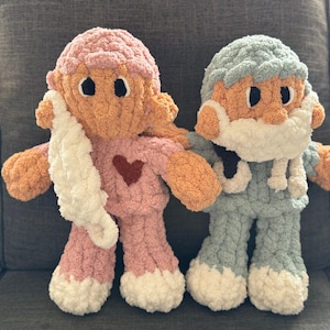 Op de afbeelding: Twee gehaakte pluche speelgoedjes, een in roze met een witte sjaal en een rood hart, en de andere in blauw met een witte baard.