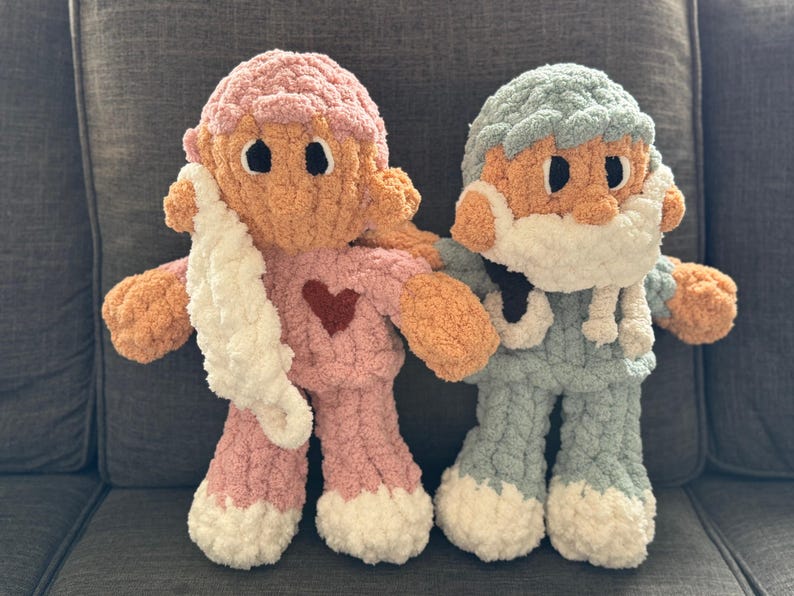 Op de afbeelding: Twee gehaakte pluche speelgoedjes, een in roze met een witte sjaal en een rood hart, en de andere in blauw met een witte baard.