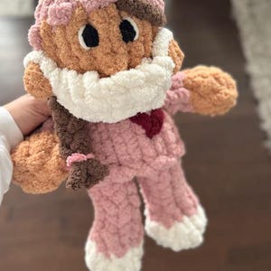 Op de afbeelding: Een gehaakte pop van roze en bruin garen, met een witte gehaakte muts en een roze en witte gehaakte outfit. De pop heeft bruin garenhaar en een rood hart op de borst.