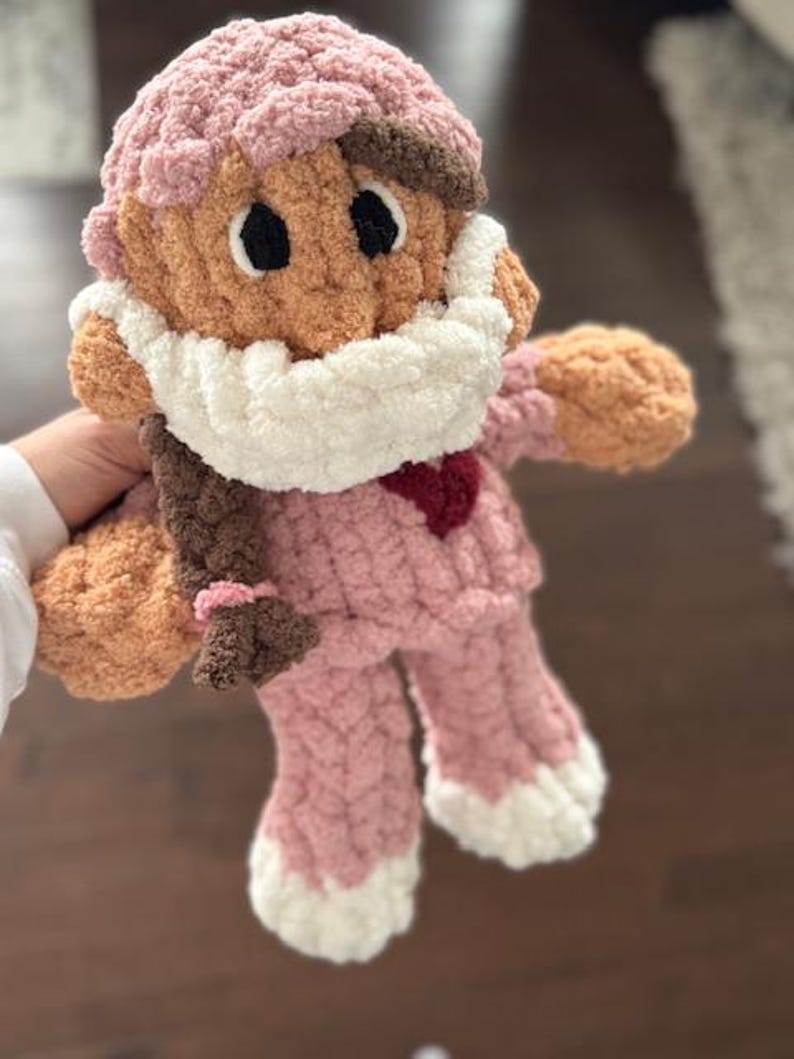 Op de afbeelding: Een gehaakte pop van roze en bruin garen, met een witte gehaakte muts en een roze en witte gehaakte outfit. De pop heeft bruin garenhaar en een rood hart op de borst.