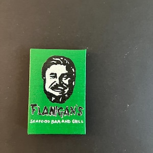 May include: A rectangular green canvas featuring a black and white graphic illustration of a man's face. Below the face, the text reads "FLANIGAN'S" in a bold, stylized font, followed by the words "SEAFOOD BAR AND GRILL" in a smaller, plain font.