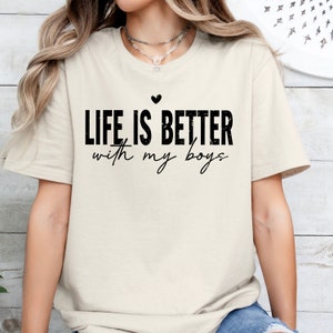 May include: A beige t-shirt with the text "LIFE IS BETTER with my boys" in black with a small heart above the text.