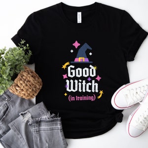 May include: Black t-shirt with a graphic of a witch's hat and the text "Good Witch (in training)" in pink and yellow lettering.