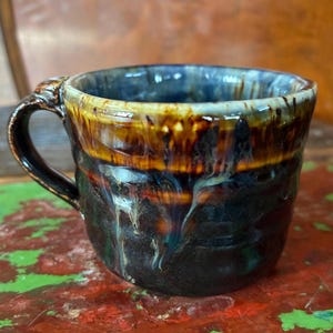May include: A ceramic mug featuring a dark, textured base that transitions into a glossy, amber-brown glaze near the rim. The surface displays vertical, dripped glaze patterns in shades of blue, white, and brown, with a curved handle on the side.