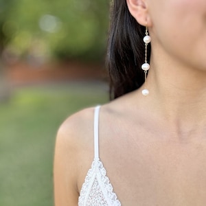 May include: Elegant gold-tone earrings featuring three luminous white pearls suspended on delicate chains. The earrings dangle gracefully, showcasing a classic and sophisticated design. Perfect for adding a touch of refined beauty to any outfit.