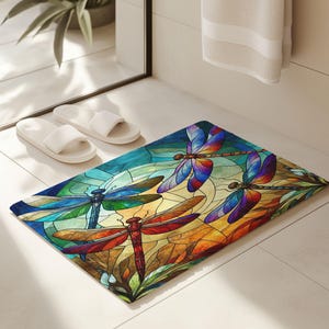 May include: A rectangular bath mat featuring a stained-glass design with four dragonflies. The dragonflies have colorful wings in shades of blue, red, and yellow. The background is a mix of blue, green, and orange.