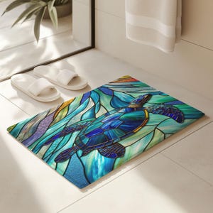 Peut inclure: Un tapis de bain rectangulaire avec un motif de vitrail représentant une tortue de mer dans des tons bleus et verts. La tortue présente un motif de mosaïque détaillé. Le tapis est posé sur un sol carrelé blanc, avec des chaussons blancs et une serviette.