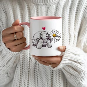 Puede incluir: Taza de cer&aacute;mica blanca con borde rosa. La taza presenta una ilustraci&oacute;n en blanco y negro de una tortuga sosteniendo una margarita en su boca. Un peque&ntilde;o coraz&oacute;n rojo est&aacute; por encima de la tortuga.