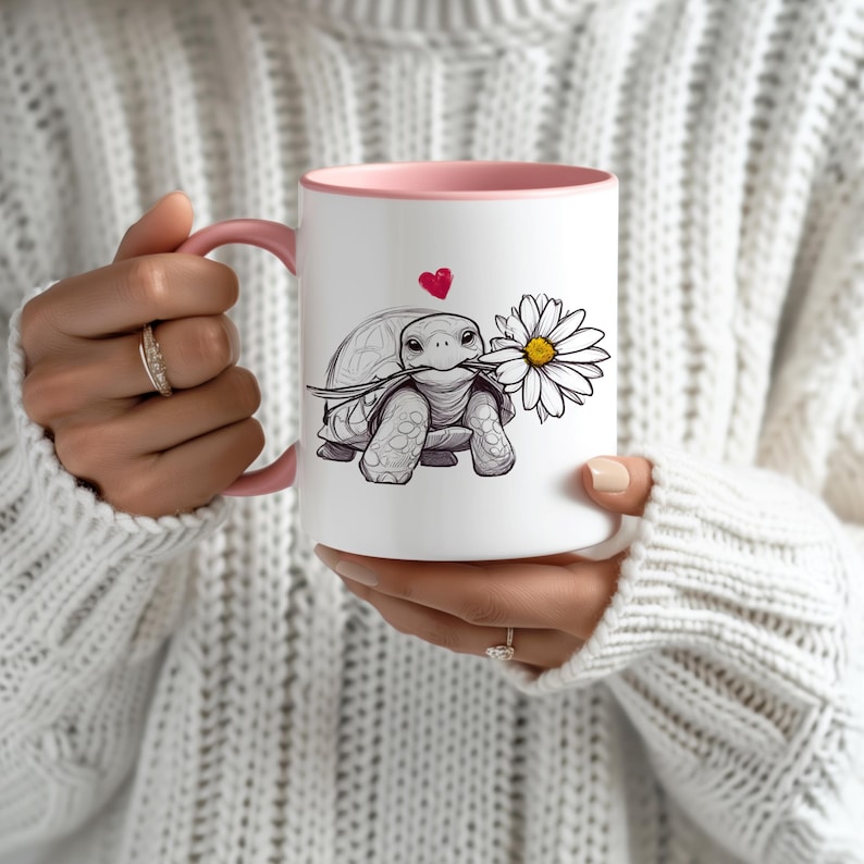 Puede incluir: Taza de cer&aacute;mica blanca con borde rosa. La taza presenta una ilustraci&oacute;n en blanco y negro de una tortuga sosteniendo una margarita en su boca. Un peque&ntilde;o coraz&oacute;n rojo est&aacute; por encima de la tortuga.