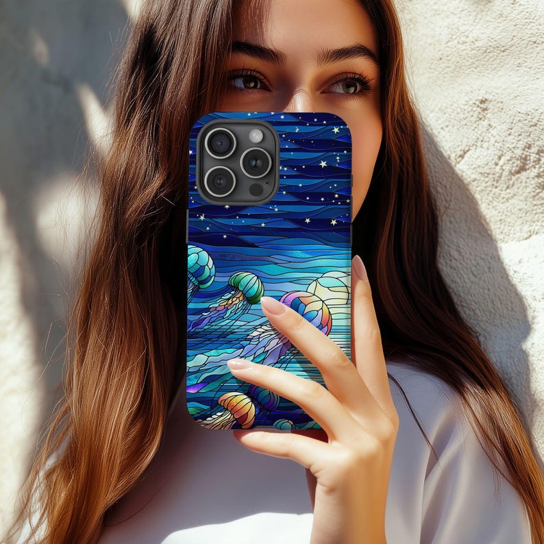 May include: A blue phone case featuring a stained-glass style design of jellyfish in the ocean. The case has a dark blue background with white stars and horizontal lines. The jellyfish are in shades of blue, green, yellow, and purple.