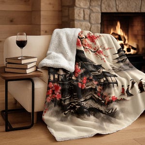 May include: A cream-coloured throw blanket with a Japanese-inspired design featuring black buildings, red flowers, and figures. The blanket is draped over a beige chair with a white sherpa lining. A glass of red wine and books sit on a side table.