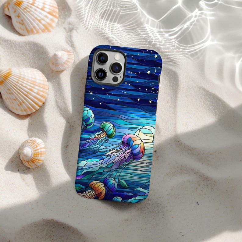 May include: A blue iPhone case featuring a stained-glass style design of jellyfish in shades of blue, purple, and orange. The case has a dark blue background with white stars and a wavy pattern. The case is lying on a sandy surface with seashells.