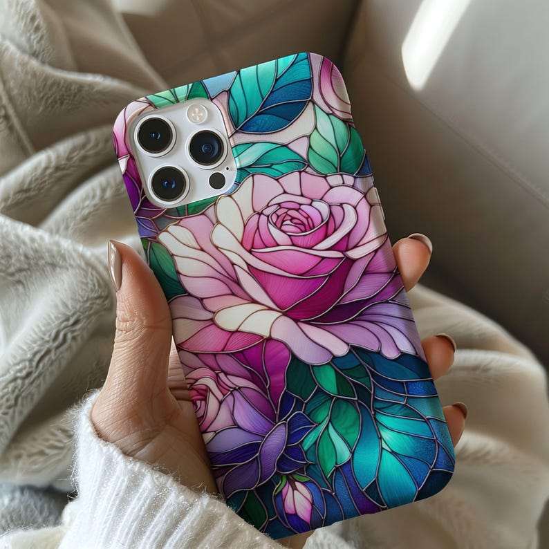 May include: A smartphone case featuring a stained-glass design of a pink rose with green and blue leaves. The case has a glossy finish and a protective bumper. The phone case is held in a person's hand.
