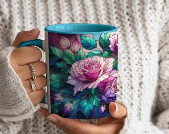 Può includere: Una tazza in ceramica bianca con interno e manico turchese, con un motivo a rosa in stile vetrata. La rosa è rosa e bianca, circondata da foglie verdi e uno sfondo blu scuro. La tazza è tenuta da una persona.