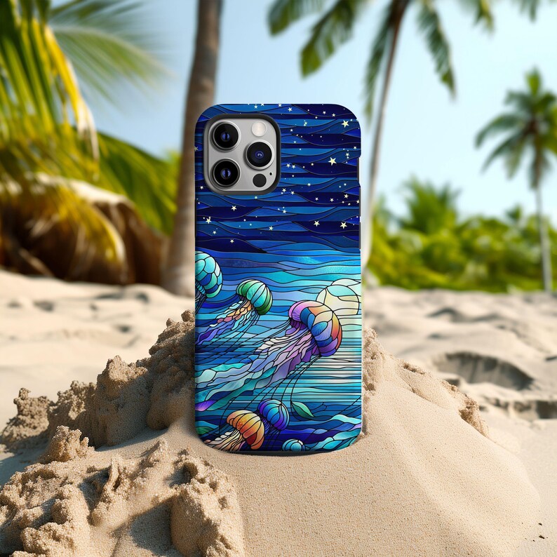 May include: A smartphone case featuring a colorful stained-glass design of jellyfish swimming in the ocean under a starry night sky. The case has a dark blue background with shades of blue, purple, orange, and green. The case is resting on a sandy beach.