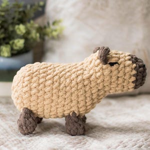 May include: A crocheted stuffed animal shaped like a capybara. The capybara is beige with brown ears and feet. It is sitting on a white, fluffy surface.