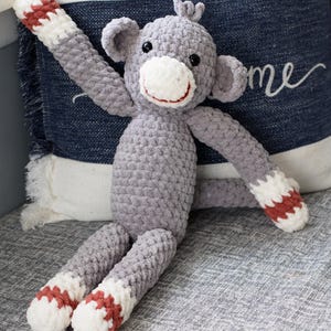 Puede incluir: Un juguete de mono de crochet gris con detalles rojos y blancos en los brazos y los pies. El mono está sentado en un sofá gris con una almohada detrás.