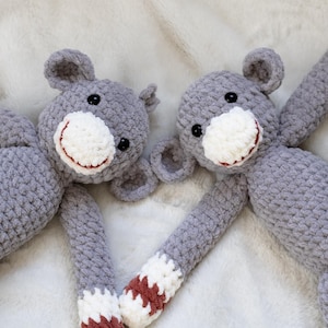 May include: Two grey crocheted monkey toys with white faces and red and white striped arms. The monkeys are lying on a white fluffy surface.