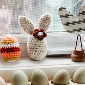 May include: A crocheted bunny with a brown flower on its head, a striped crocheted egg, and a wooden rooster figurine on a white windowsill. There are also six plain eggs in an egg carton.