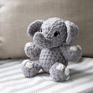 Puede incluir: Un peluche de elefante gris tejido a ganchillo con detalles blancos en las patas. El elefante tiene orejas grandes, una trompa y ojos negros. Está sentado sobre una superficie a rayas con una almohada de color neutro en el fondo. A la izquierda hay una pequeña planta en maceta.