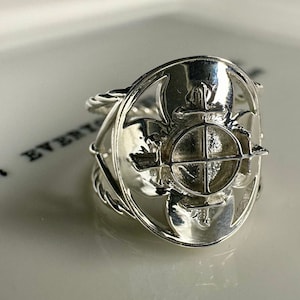 May include: A silver ring with a detailed design featuring a circular center with a cross and a flower-like pattern around it.