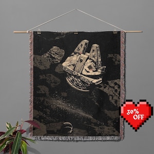 Puede incluir: Un tapiz en blanco y negro con una nave espacial volando por el espacio. La nave espacial es un Millennium Falcon, una nave espacial popular de la franquicia Star Wars. El tapiz tiene un borde con flecos y está colgado en una pared.