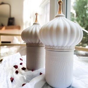 May include: Two white ceramic candle jars with gold lids shaped like domes. The jars have a textured design and are sitting on a white fabric surface.