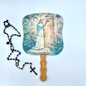 Puede incluir: Un abanico vintage con una imagen religiosa de Jesús llamando a una puerta. El abanico tiene un mango de madera y un diseño de papel en colores azul, verde y blanco. También se ve un rosario azul con una cruz de plata.