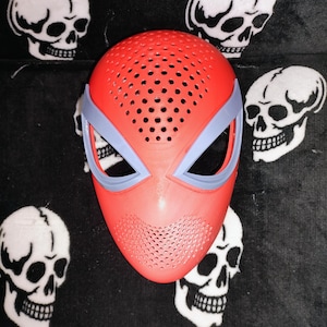 May include: A red and light blue mask with a perforated design, similar to a superhero mask. The mask has eye openings and a mouth area with small holes. The background features white skull designs on a black surface.