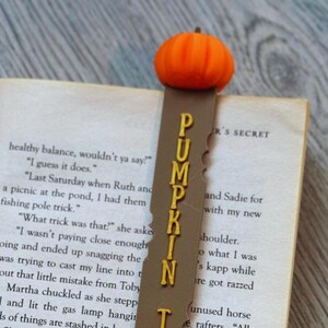 Könnte beinhalten: Ein braunes Lesezeichen mit einem Kürbisdesign und den Worten "Pumpkin Time" in goldenen Buchstaben. Das Lesezeichen befindet sich auf einem Buch mit dem sichtbaren Text "R's Secret".