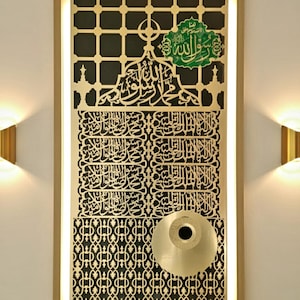 May include: Framed wall art featuring gold-coloured Arabic calligraphy and geometric patterns. The artwork includes a gold-coloured circular element and a green accent with text. The frame is light brown.