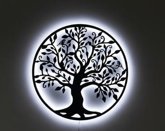 May include: A black metal tree of life wall art with a circular design. The tree features intricate branches, leaves, and two birds. The art is illuminated by a white LED light, creating a halo effect. A power cord is visible.