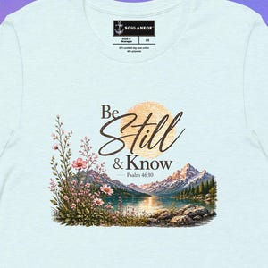 May include: Pale blue t-shirt featuring a scenic graphic of a lake and mountains, with the text "Be Still & Know" from Psalm 46:10. The design incorporates floral details and a sun. The shirt has a round neck and short sleeves.