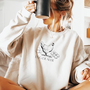 May include: Beige sweatshirt with a black graphic of a hen, eggs, and the text "EGG DEALER." The sweatshirt is long-sleeved and has a crew neck. The person is holding a black mug.