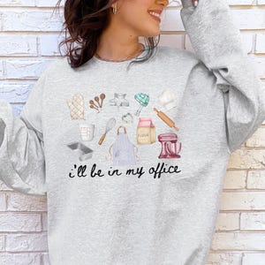 May include: A light gray crewneck sweatshirt featuring a graphic print of various baking tools including a whisk, rolling pin, apron, and stand mixer, with the black text "i'll be in my office" printed underneath the collection of kitchen items.