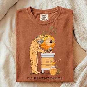 May include: Brown t-shirt with a graphic of a beekeeper in a yellow suit tending to a beehive. A honey pot and the text "I'll be in my office" are also featured. The shirt is made of Comfort Colors material.