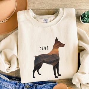 May include: Cream-colored sweatshirt featuring a silhouette of a dog with a mountain landscape design. The word "COCO" is printed above the dog. The sweatshirt is paired with blue jeans, a brown hat, and sandals.