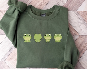 May include: An olive green crewneck sweatshirt featuring a graphic print of four light green frogs in different poses. The frogs are arranged horizontally across the center of the chest, showing various expressions and postures in a simple cartoon style.