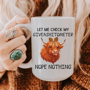 May include: White ceramic mug with the text "LET ME CHECK MY GIVEASHITOMETER" and "NOPE NOTHING". The mug features a cartoon illustration of a Highland cow with a brown bow and its tongue sticking out.
