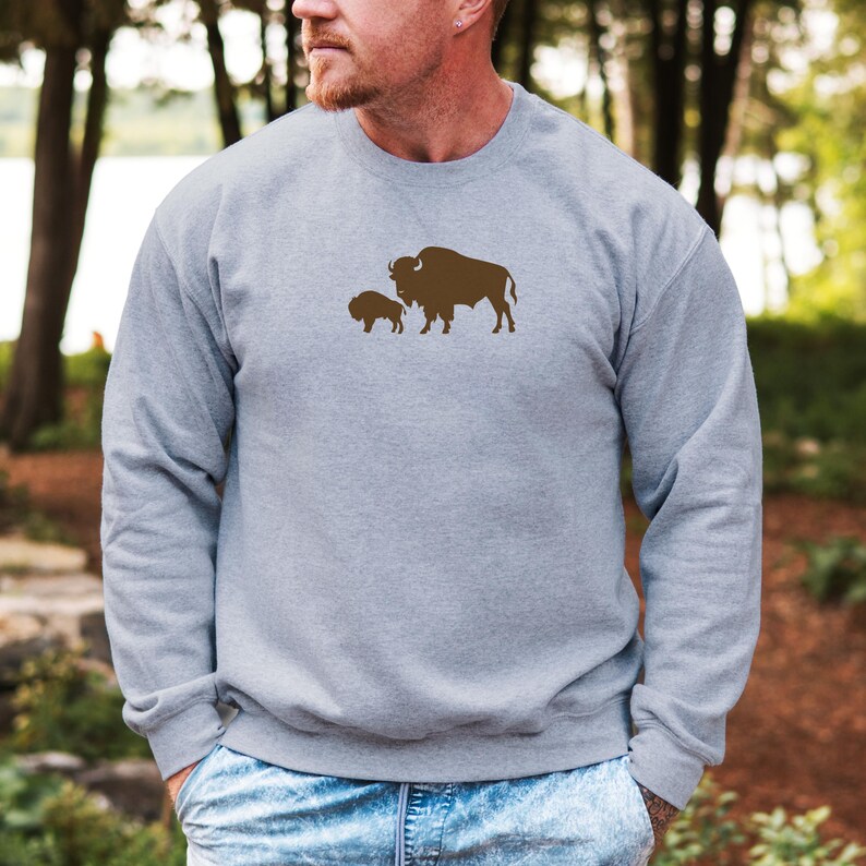 May include: A heather grey crewneck sweatshirt featuring a brown graphic print of a large bison standing next to a smaller bison calf centred on the chest. The garment has long sleeves, a ribbed neckline, and a standard fit construction.