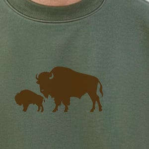 May include: An olive green crewneck sweatshirt featuring a dark brown graphic print of an adult bison standing next to a smaller bison calf. The sweatshirt has a ribbed collar and long sleeves, showcasing a simple, clean design on the front.