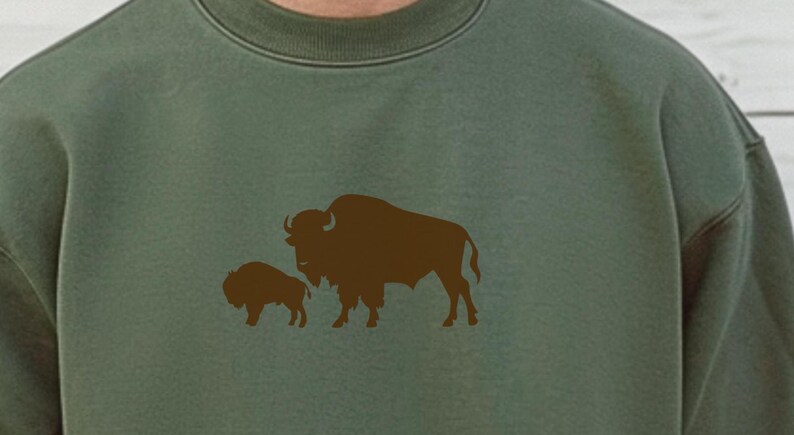 May include: An olive green crewneck sweatshirt featuring a dark brown graphic print of an adult bison standing next to a smaller bison calf. The sweatshirt has a ribbed collar and long sleeves, showcasing a simple, clean design on the front.