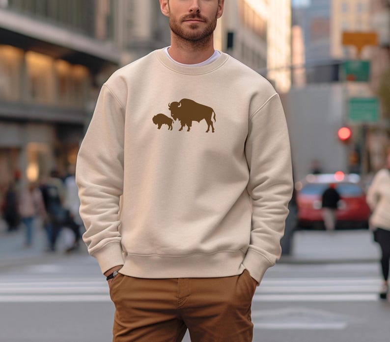May include: A cream-colored sweatshirt featuring a brown bison graphic. The crew neck sweatshirt has a relaxed fit and is made of a soft material. The bison graphic includes a larger bison and a smaller bison. The sweatshirt is paired with brown pants.
