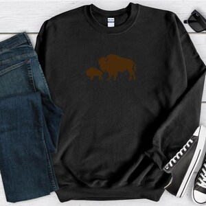May include: A black long-sleeve crewneck sweatshirt featuring a brown graphic of a large bison and a smaller bison calf centred on the chest. The garment has a ribbed collar, cuffs, and hem, with a Gildan brand label visible at the inner neckline.